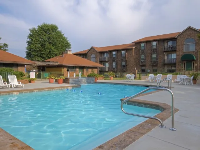 Large outdoor pool surrounded by a concrete deck with lounge chairs, set in front of a three-story apartment building with red tile roofing.