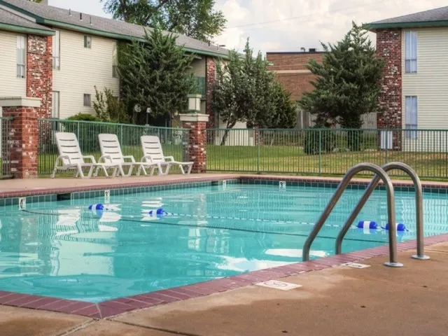 Outdoor swimming pool with clear water, surrounded by concrete decking, white lounge chairs, a black chain-link fence, and mature trees in the background.