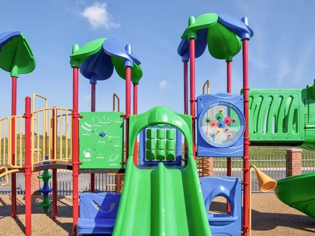 Colorful outdoor playground at Embassy Apartments in Ozark, Missouri, featuring green and purple plastic structures with slides, climbing panels, and a protective rubber surface.