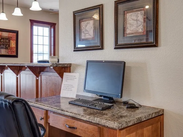 Business center interior at Embassy Apartments in Ozark, Missouri, showcasing a granite-topped desk with a desktop computer, built-in wood cabinetry, framed artwork on neutral walls, and daylight streaming through windows.