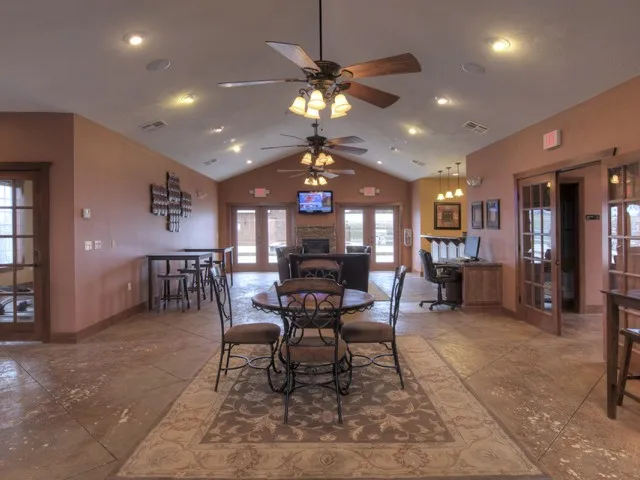 Clubhouse lounge at Embassy Apartments in Ozark, Missouri, with round tables and chairs on a patterned rug, wood-look flooring, ceiling fans, recessed lighting, and decorative shelving along the walls.