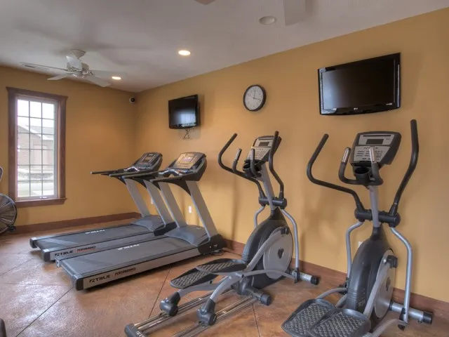 On-site fitness center at Embassy Apartments in Ozark, Missouri, featuring treadmills and elliptical machines lined up against warm-colored walls beneath wall-mounted televisions.