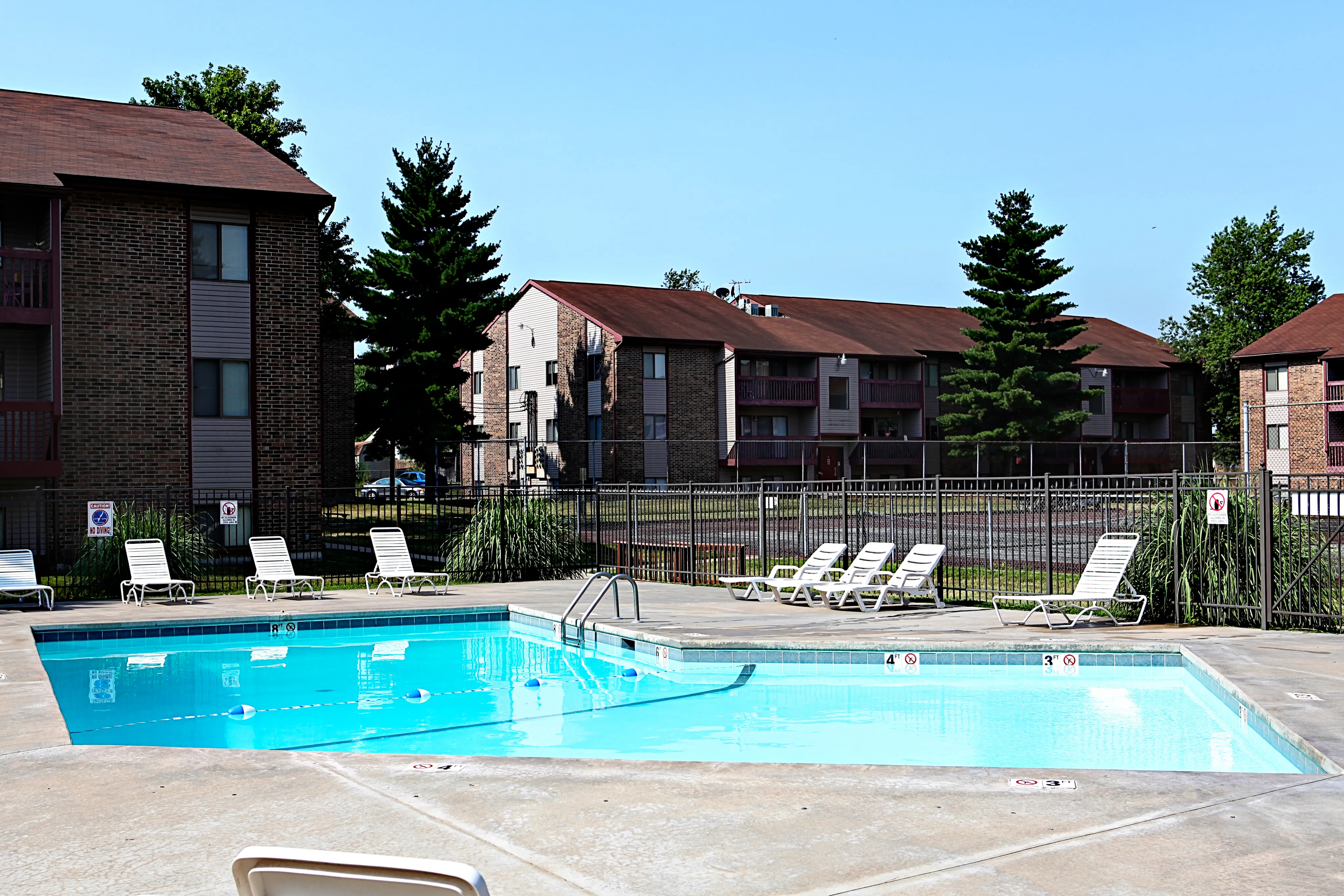 Outdoor swimming pool with a concrete deck, lounge chairs, and surrounding fence, set against a backdrop of two-story apartment buildings and trees.