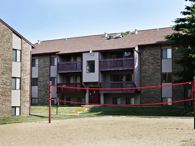 Sand volleyball court with a red net located between two brick apartment buildings with balconies and sloped roofs.