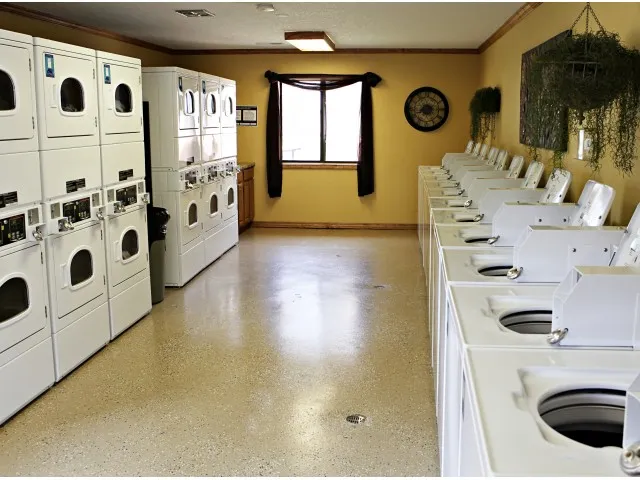 Spacious community laundry room with rows of top-loading washers on one side and stacked front-loading dryers on the other, featuring warm yellow walls and natural light from a window.