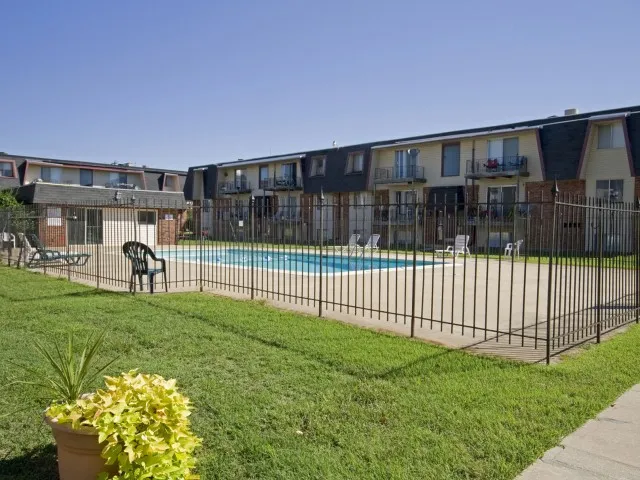 Gated swimming pool area at Rosewood Village Apartments with surrounding lawn and two-story buildings in the background.