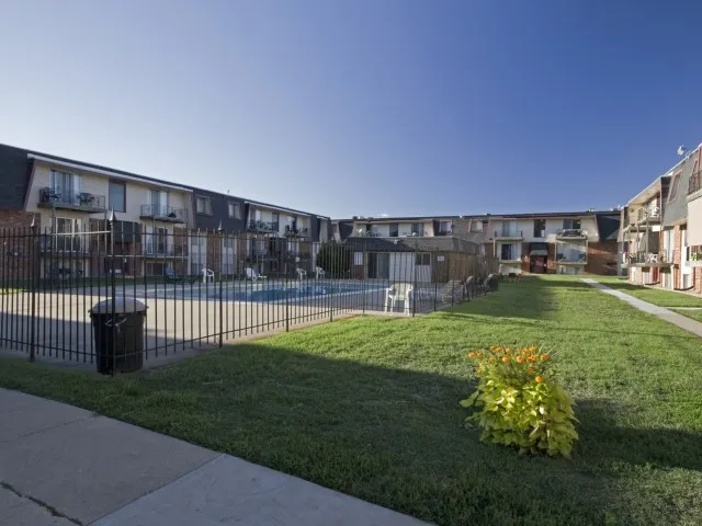 Wide view of the fenced pool at Rosewood Village Apartments, showing expansive green space and apartment exteriors on a clear day.