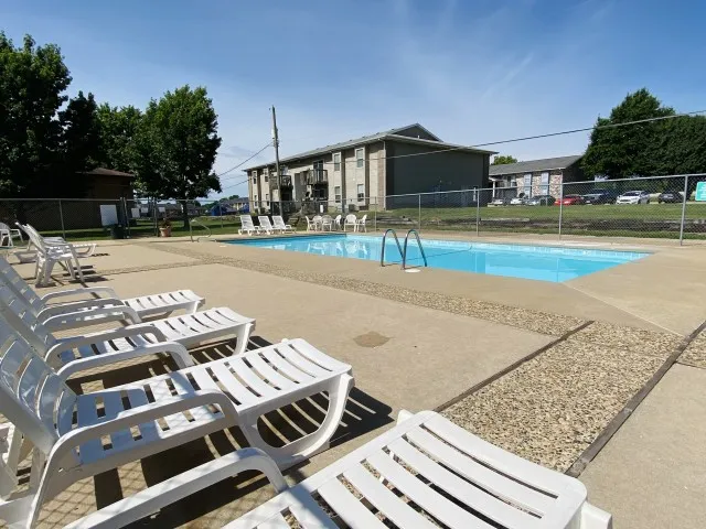 Side view of the pool area with multiple lounge chairs and buildings in the background under a bright sky.