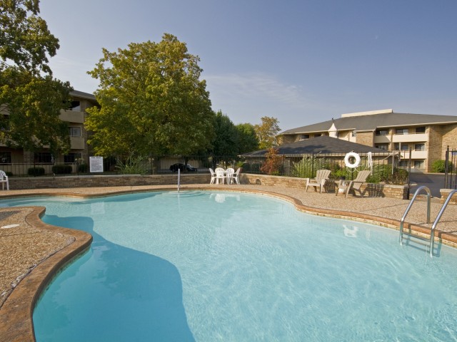 Outdoor swimming pool in a sunny courtyard, surrounded by trees and buildings. Lounge chairs and a life preserver are visible, creating a relaxed atmosphere.