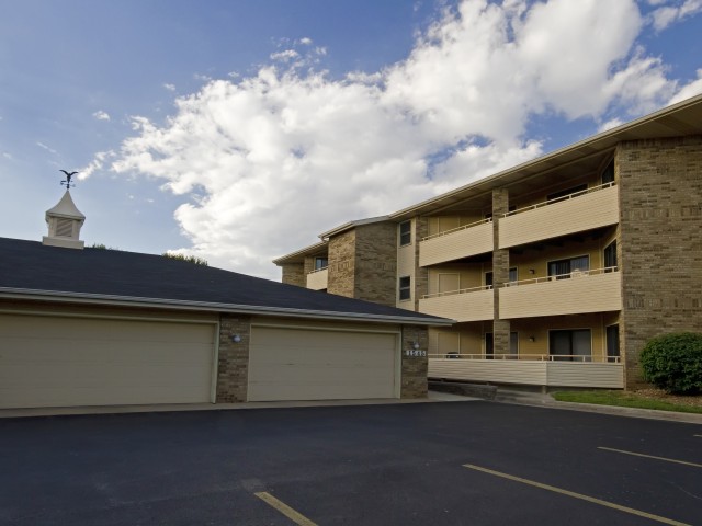 Two beige buildings under a partly cloudy sky, featuring a three-story apartment complex and a one-story garage with stone accents. Calm, suburban setting.