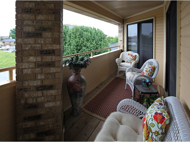 A cozy veranda with wicker chairs and floral cushions on a woven rug. A large vase and potted plants sit near a brick wall, creating a tranquil ambiance.