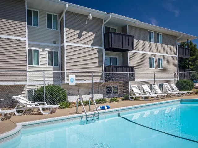Outdoor swimming pool at Southwinds Apartments in Springfield, MO, surrounded by white lounge chairs and a safety fence. The three-story apartment building features beige siding and dark balcony railings under a clear blue sky.