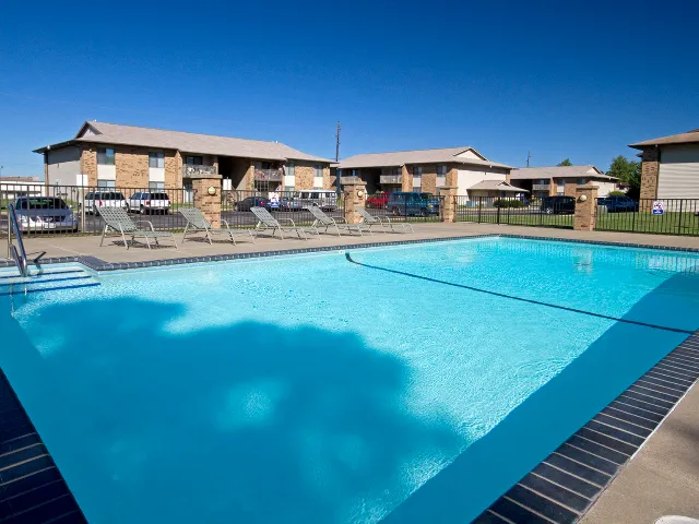 Outdoor resort-style swimming pool at English Village Apartments in Springfield, Missouri, with crystal-blue water, a concrete deck lined with lounge chairs, and one- and two-story tan brick apartment buildings and manicured landscaping under a clear blue sky.