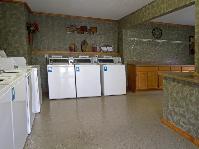 Community laundry room at English Village Apartments in Springfield, Missouri, featuring a row of white top-loading washing machines against green patterned walls, a speckled epoxy floor, and wood-tone upper cabinets for storage.