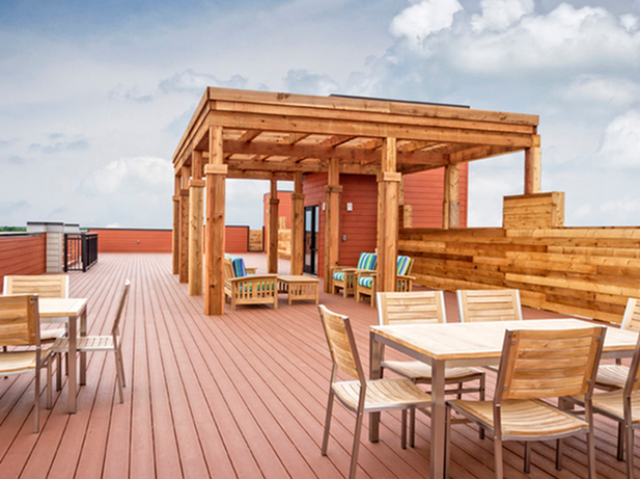 Spacious rooftop deck with wooden pergola, outdoor seating, and dining tables set on red composite decking under a partly cloudy sky.