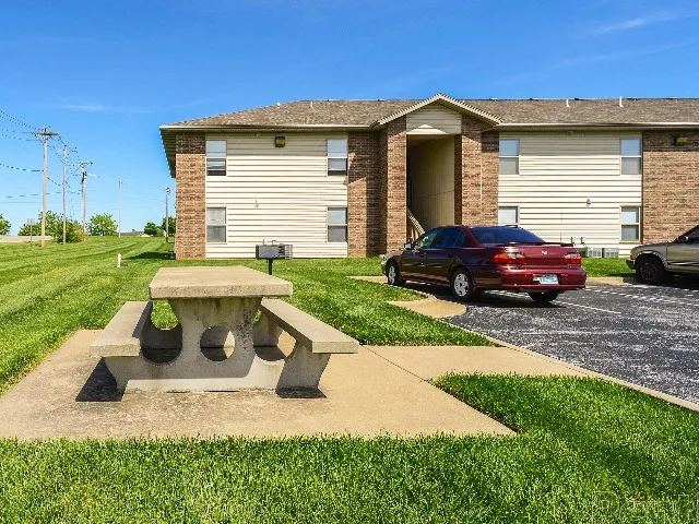 Concrete picnic table on a paved pad in front of a Huntley Ridge East apartment building, with a red car parked nearby and a well-kept lawn on a sunny day.