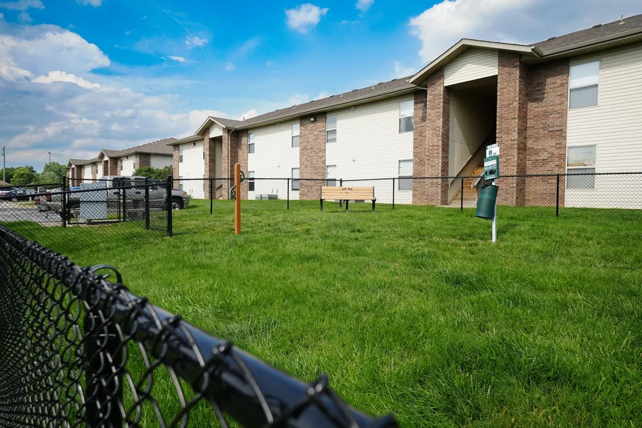 Wide-angle view of a fenced-in dog park area at Huntley Ridge East Apartments in Springfield, MO, showing a green pet waste station, wooden bench, and dog leash post, all set against a backdrop of brick apartment buildings and blue sky.