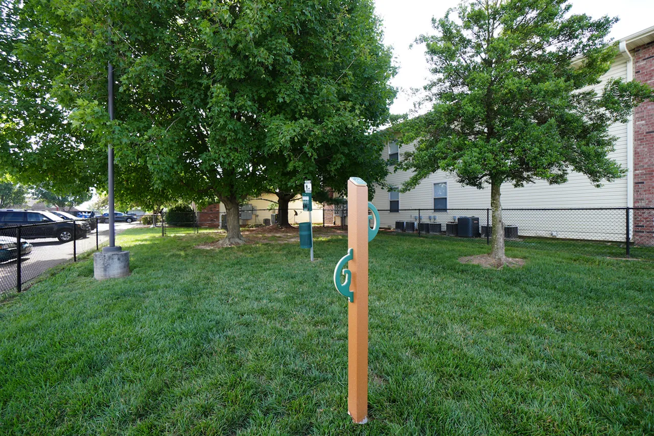 Spacious, grassy dog park at Huntley Ridge Apartments with mature trees, a fenced perimeter, pet waste station, and a pet play post for leashing or tethering. Apartment buildings and parking lot visible in the background.