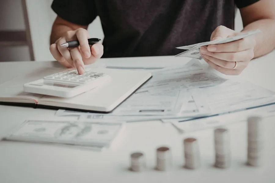 Focused individual calculating expenses with a calculator, surrounded by financial documents and stacks of coins.