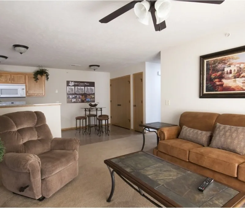 A cozy living room featuring a brown sofa, armchair, and glass coffee table, with a ceiling fan overhead. An adjoining kitchen and dining area are visible.