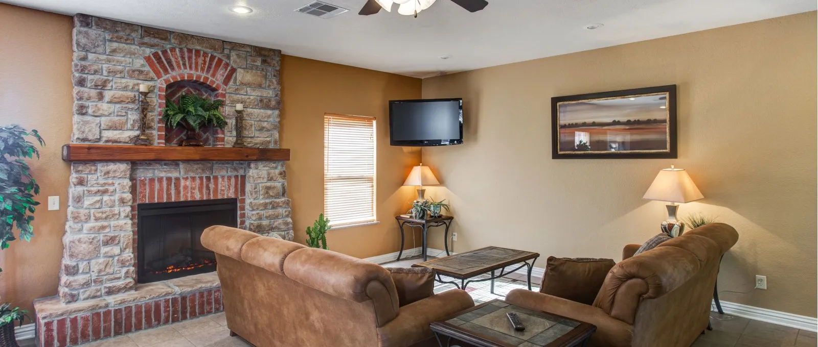 Clubhouse with a cozy ambiance, featuring a brown leather couch, stone fireplace, wall-mounted TV, and a small table with a lamp and plant.