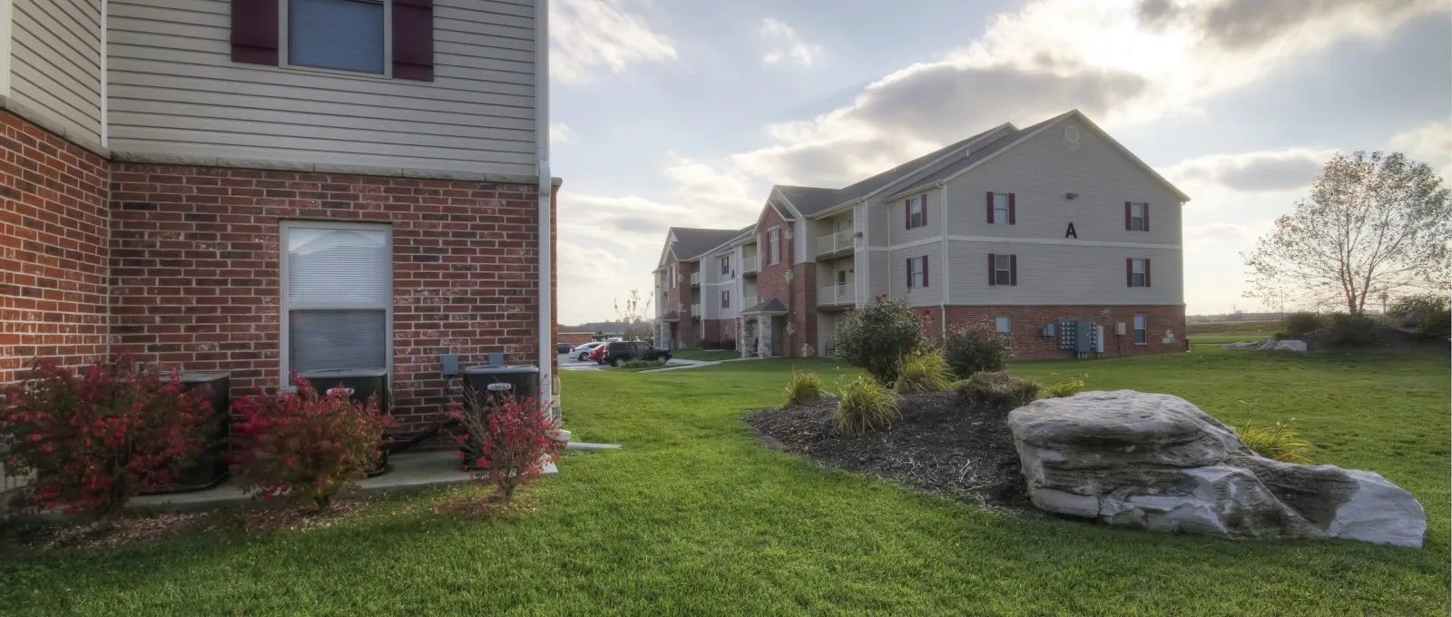 Apartment buildings under a cloudy sky with the sun peeking through. A grassy courtyard and neatly landscaped areas create a calm suburban setting.