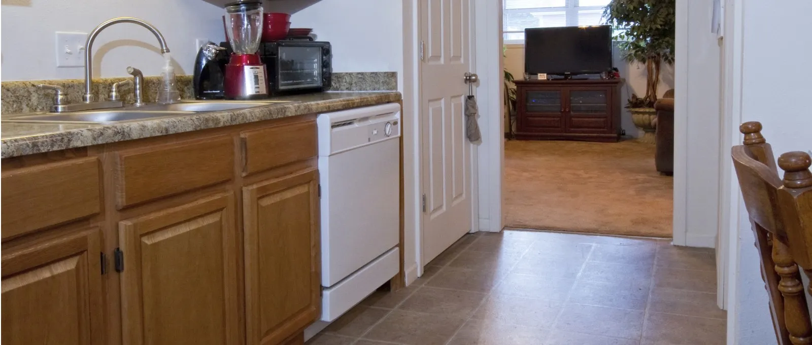 Kitchen with wood cabinets, a white dishwasher, and countertop appliances. Doorway leads to a cozy living room with a TV stand and neutral decor.