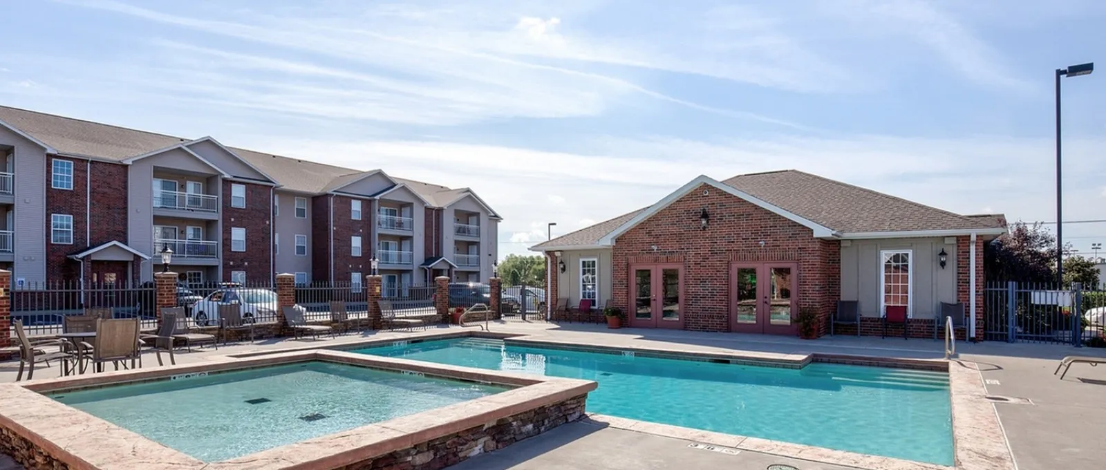 Resort-style outdoor swimming pool at Embassy Apartments in Ozark, Missouri, featuring crystal-blue water edged with stone coping, a brick clubhouse building to the right, and a multi-story red-brick and tan apartment building in the backgr