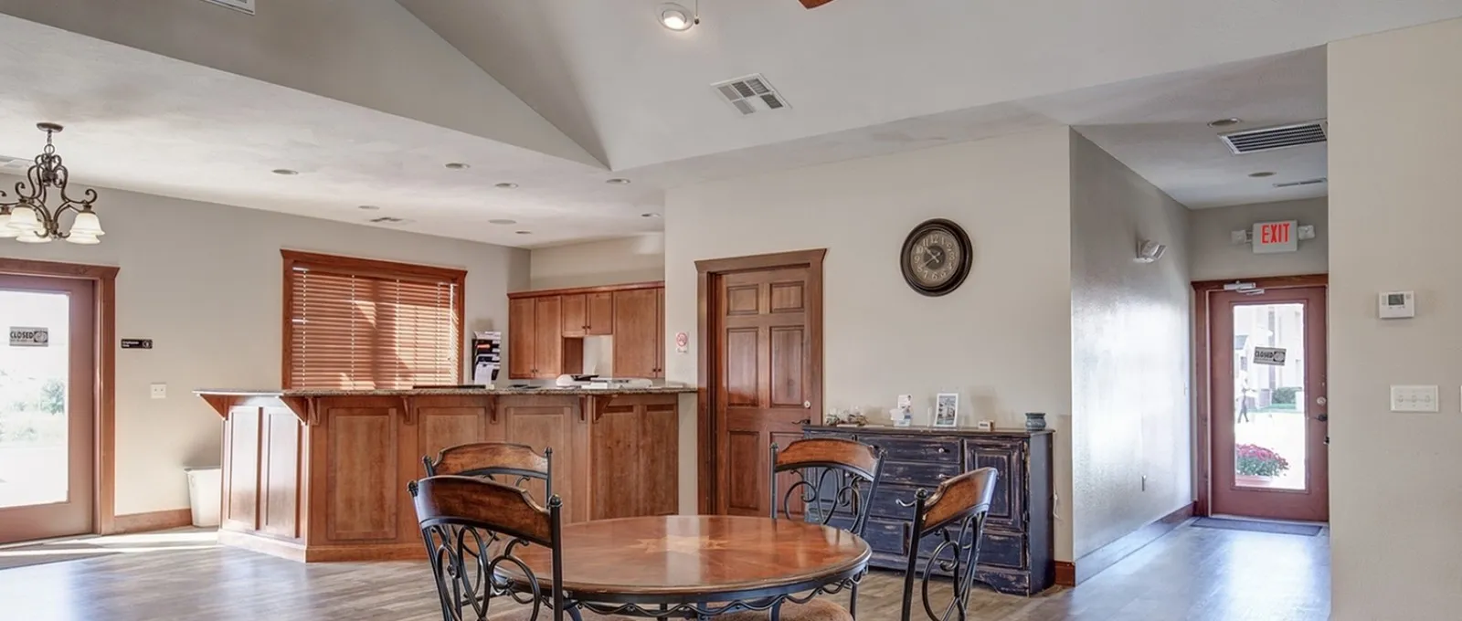 Community lounge at Embassy Apartments in Ozark, Missouri, featuring a round wood table with four metal-frame chairs, a built-in wood-tone kitchenette with granite countertop and overhead cabinets, all under recessed ceiling lights against