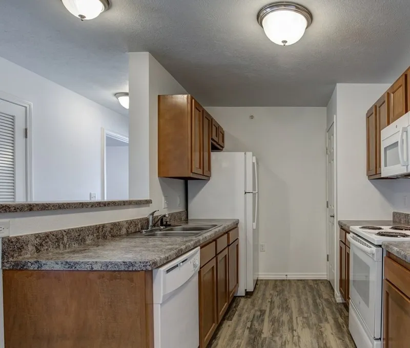 Model apartment kitchen at Embassy Apartments in Ozark, Missouri, featuring dark wood cabinets, stainless steel electric range and over-range microwave, granite countertops with a breakfast bar and three stools, and recessed lighting.