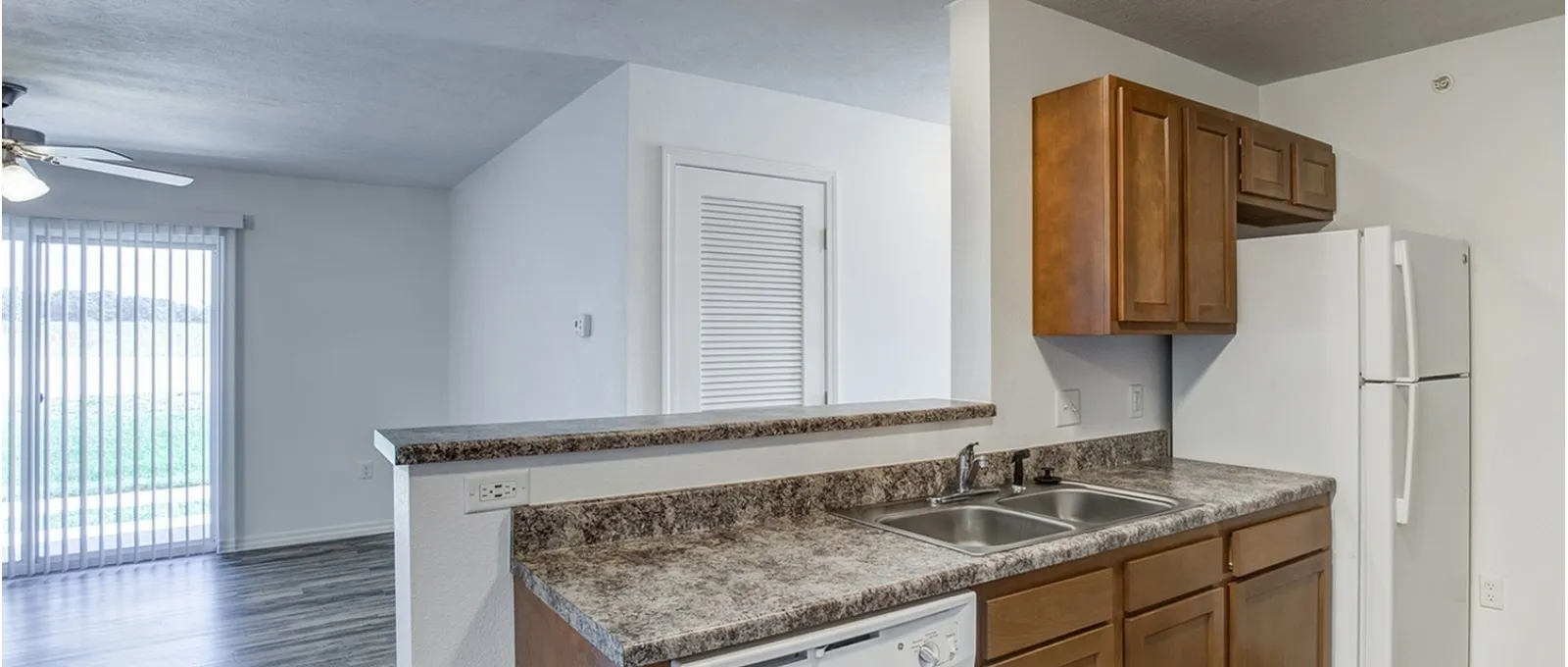 kitchen at Embassy Apartments in Ozark, Missouri, showcasing a granite countertop with undermount stainless sink, wood cabinets with brushed-nickel hardware, a tile backsplash, and a built-in microwave above the counter.