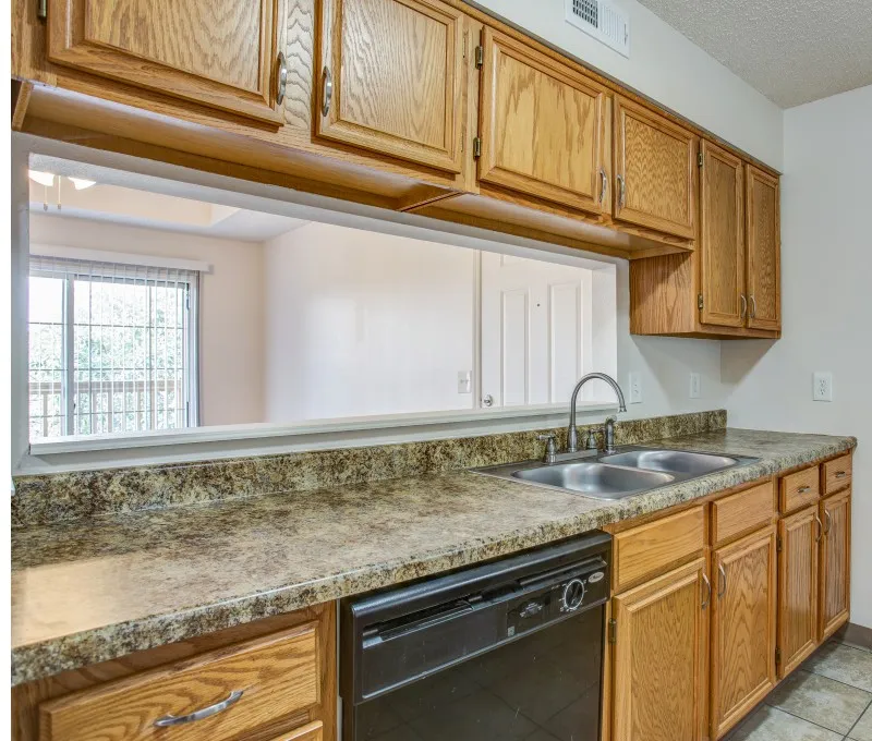 Renovated galley kitchen at Willow Creek Apartments showcasing granite-look countertops, wood cabinets, and a double sink with passthrough view.