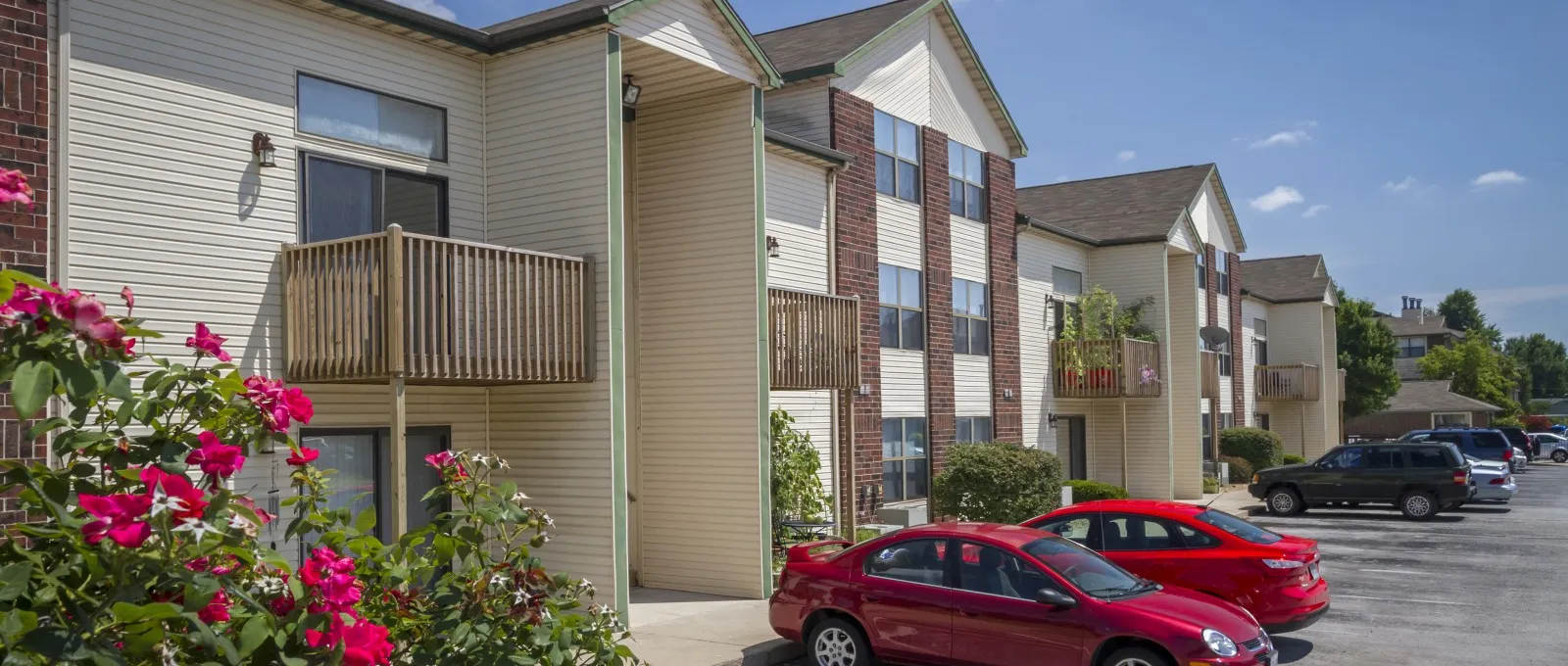 Exterior view showing the apartments’ architecture and on-site parking area with red cars, framed by blooming rose bushes and a bright blue sky.