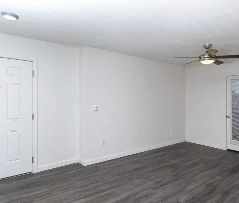 Empty room with white walls and dark wood laminate flooring. A door on the left and a ceiling fan with lights on the right create a simple, serene atmosphere.