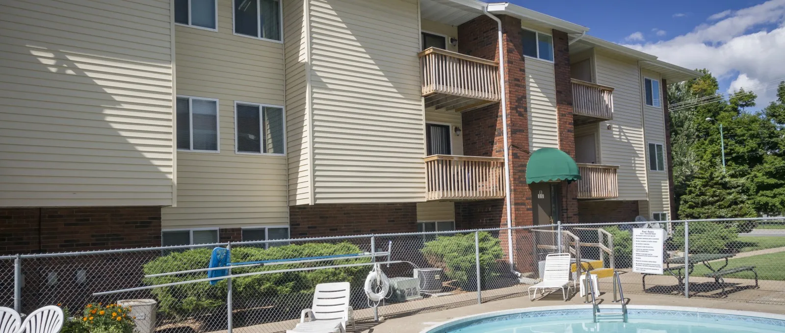 Close-up view of the outdoor swimming pool at Markwood Apartments, featuring a curved design, pool ladder, and lounge chairs within a fenced area.