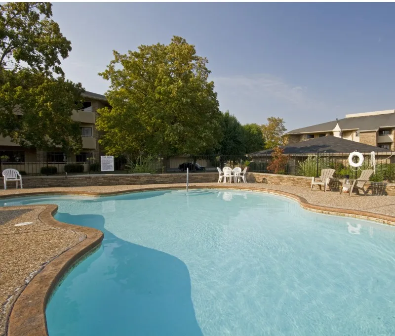Outdoor swimming pool in a sunny courtyard, surrounded by trees and buildings. Lounge chairs and a life preserver are visible, creating a relaxed atmosphere.