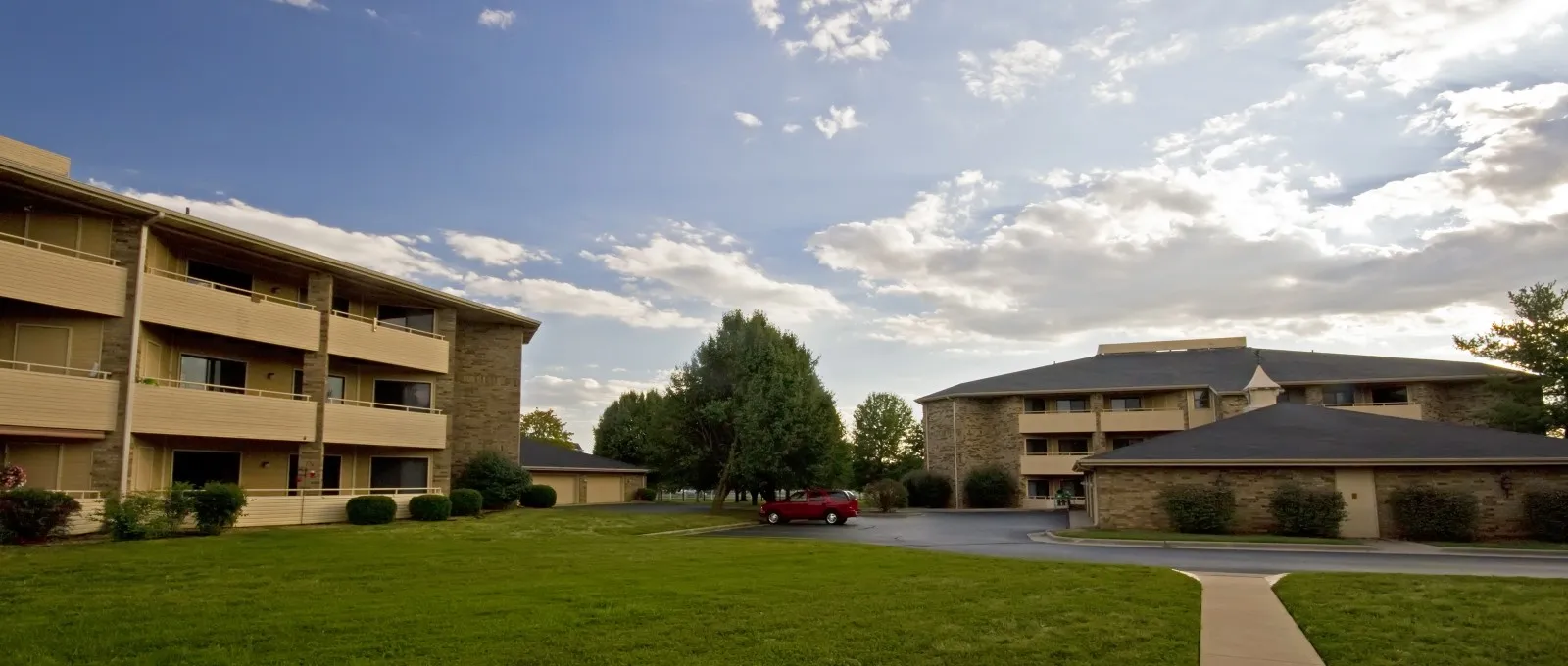 A sunny courtyard between two apartment buildings, featuring a neatly trimmed lawn and scattered clouds in the blue sky, conveys a serene atmosphere.