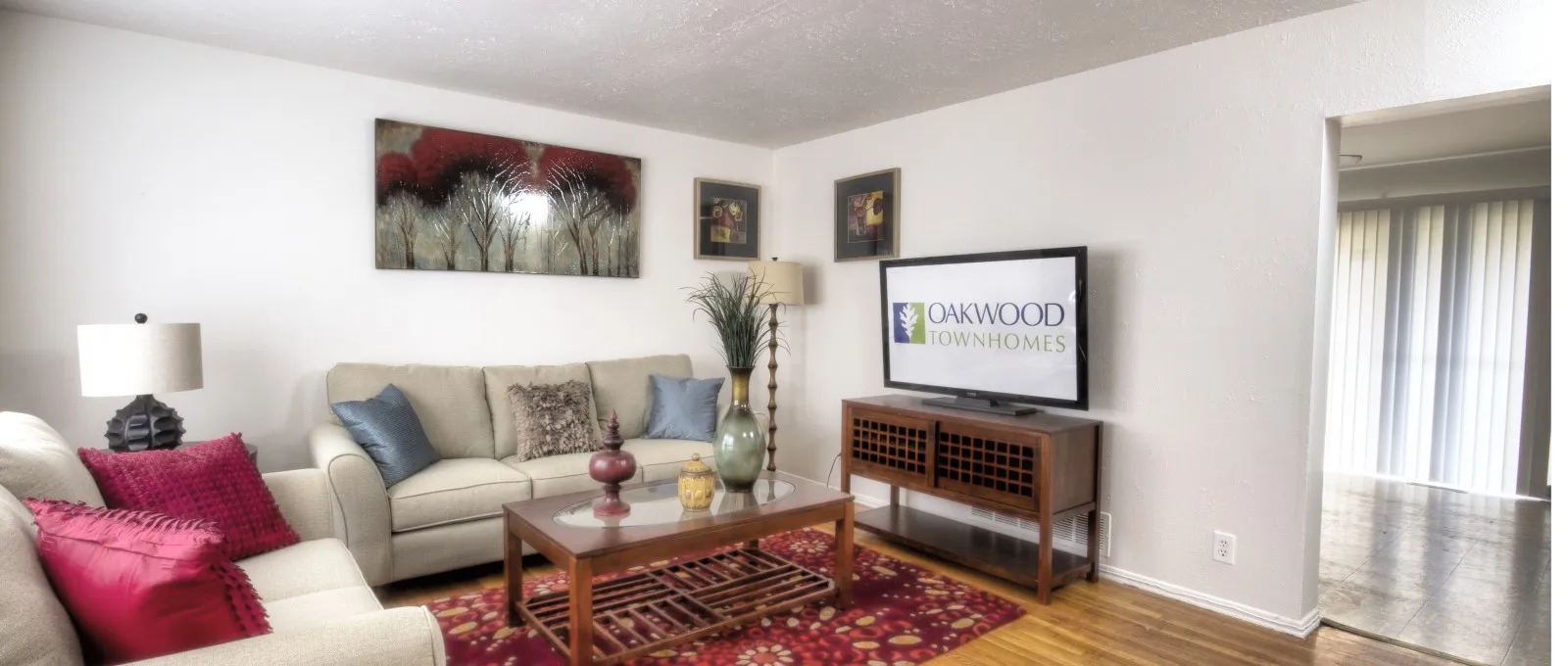 A cozy living room with a beige sofa adorned with cushions, a wooden coffee table on a red patterned rug, art on white walls, and a TV on a wooden stand.