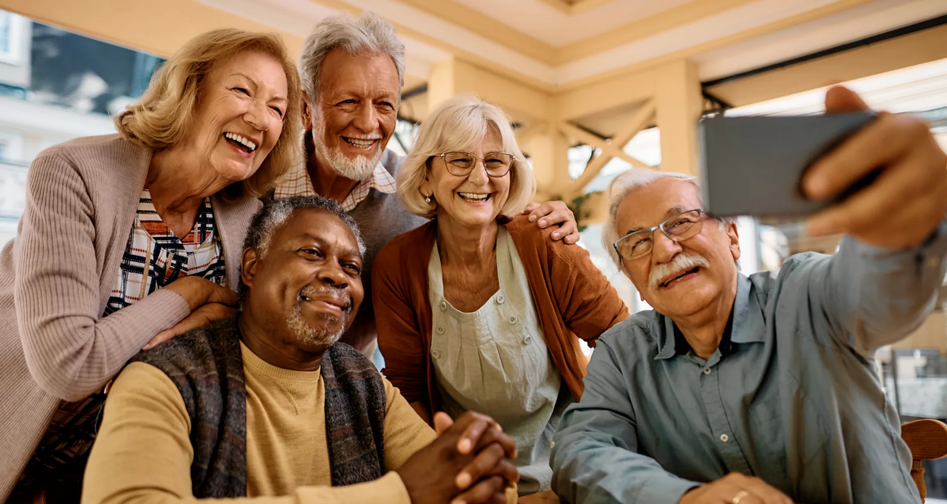 Five smiling senior adults of diverse backgrounds gathered closely indoors, leaning in toward the camera in a bright, welcoming communal space.