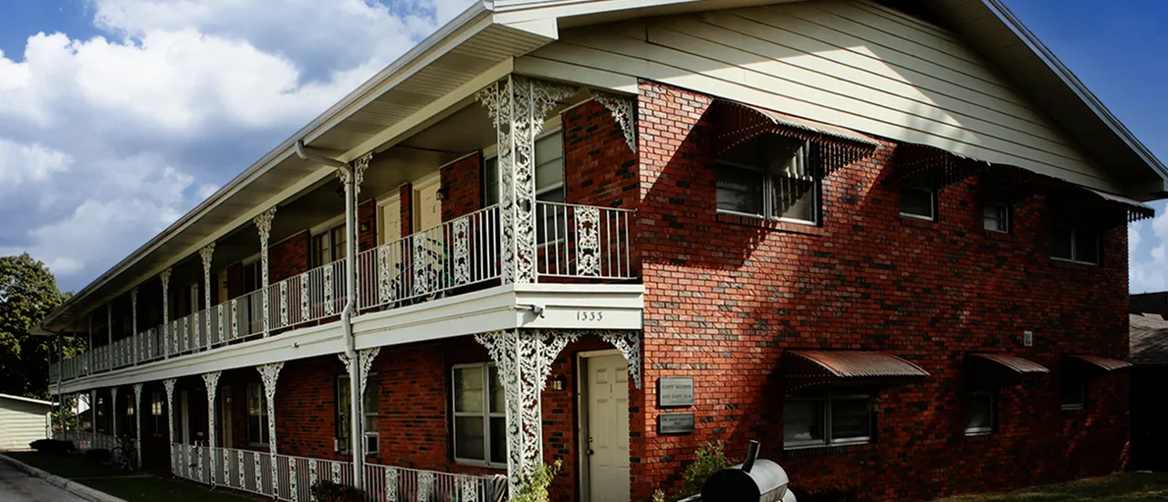 Exterior of East Manor Apartments in Springfield, Missouri—two-story red brick building with white decorative wrought-iron balcony railings, ornate trim under the eaves, and red entry doors beneath covered porches.