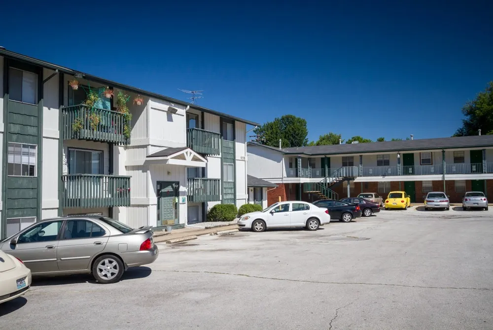 Two-story motel with white walls and balconies set against a clear blue sky. Cars are parked in the lot, suggesting a quiet, sunny day.