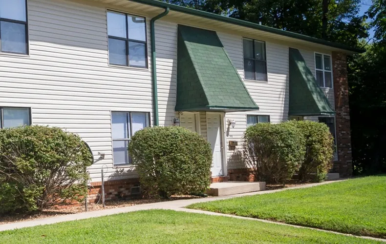 A two-story apartment building with green awnings, white siding, and neatly trimmed bushes. Bright, sunny day, creating a welcoming atmosphere.