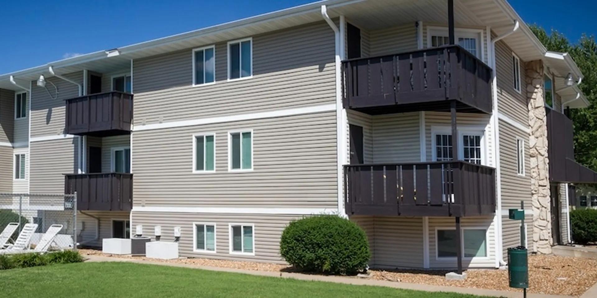 A modern two-story apartment building with gray siding, multiple windows, and black balconies, surrounded by green grass.