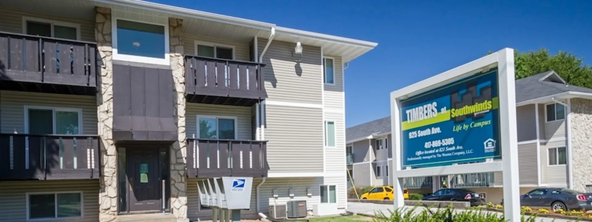 A modern apartment building features light-colored exterior, balcony, and a USPS mailbox, with bright blue skies above.