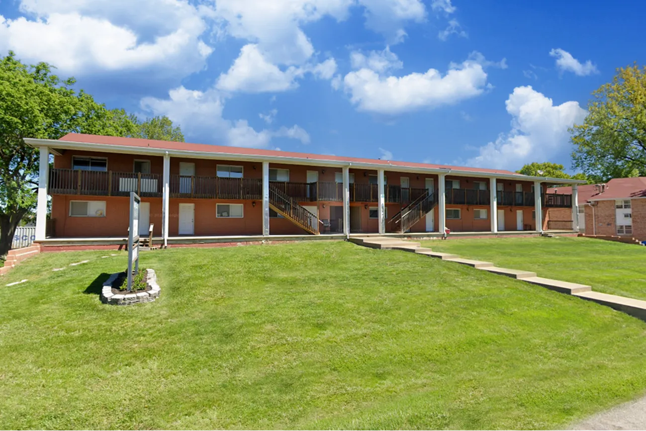 Exterior view of Glen Isle Apartments at Village Club, showing a two-story building with exterior staircases, surrounded by a large open lawn and a bright blue sky with scattered clouds.
