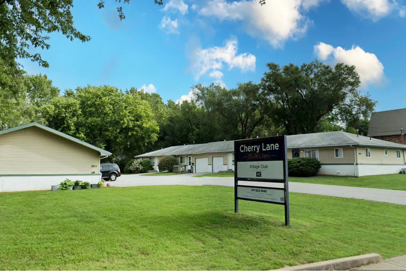 Cherry Lane Apartments entrance at Village Club featuring a community sign on a manicured lawn, with single-story buildings and tree-lined surroundings in the background.