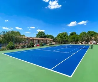 A vibrant blue tennis court under a clear sky with scattered clouds. Surrounded by trees and a few buildings, it exudes a peaceful, sunny atmosphere.