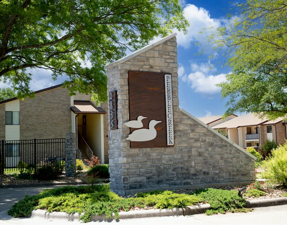 Stone monument with metal duck silhouettes and text, surrounded by greenery. Bright blue sky and apartment buildings in the background convey a welcoming tone.