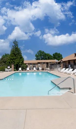 Outdoor swimming pool surrounded by lounge chairs and brick buildings, under a bright blue sky with fluffy clouds. Relaxing and inviting scene.