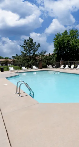 Outdoor swimming pool under a bright blue sky with scattered clouds. Surrounded by trees and lined with white lounge chairs, creating a relaxing atmosphere.
